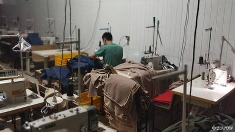 Un hombre se inclina sobre una máquina de coser industrial en una habitación llena de restos de tela y escritorios.