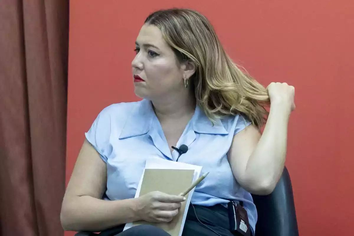 Mujer con camisa azul claro y cabello rubio sentada en una silla, sosteniendo un cuaderno y un bolígrafo, con fondo rojo.